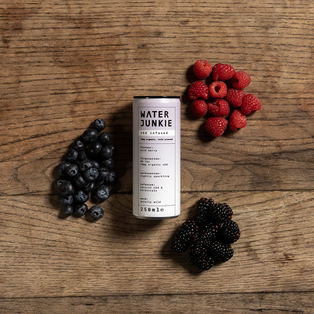 Water Junkie wild berry can laying on a wooden background surrounded by raspberries, blueberries and blackberries.