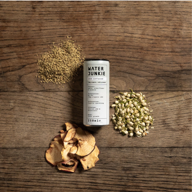 A Water Junkie apple, elderflower and jasmine can laying on a wooden table, surrounded by dried apple, elderflower and jasmine