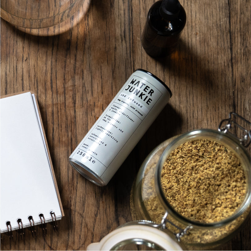 Water Junkie apple, elderflower and jasmine can laying on a wooden table next to a notepad and ingredients