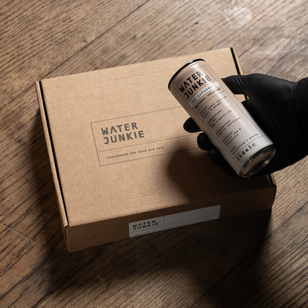A hand holding the Water Junkie white peach, passion-flower and ginger can up to the camera in front of a taster pack delivery box